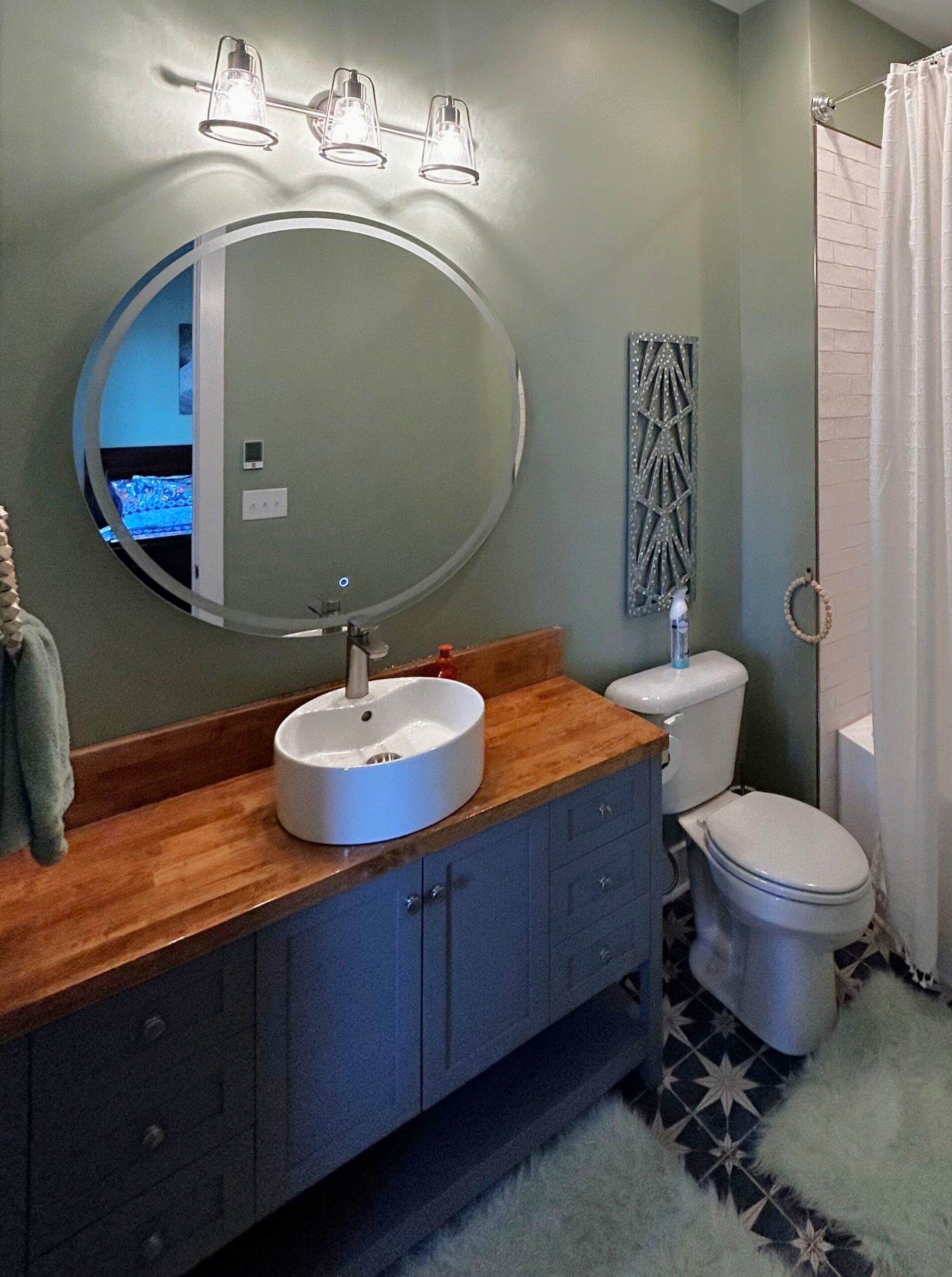Bathroom vanity with round mirror, vessel sink, wood countertop, chrome lighting, and blue cabinets.