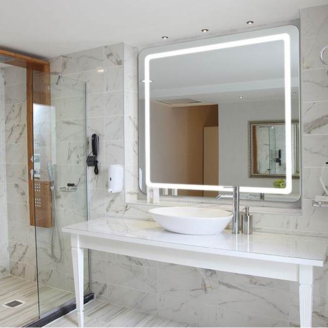 Bath and Bronze lighted bathroom mirror above a white vanity with marble walls and glass shower.