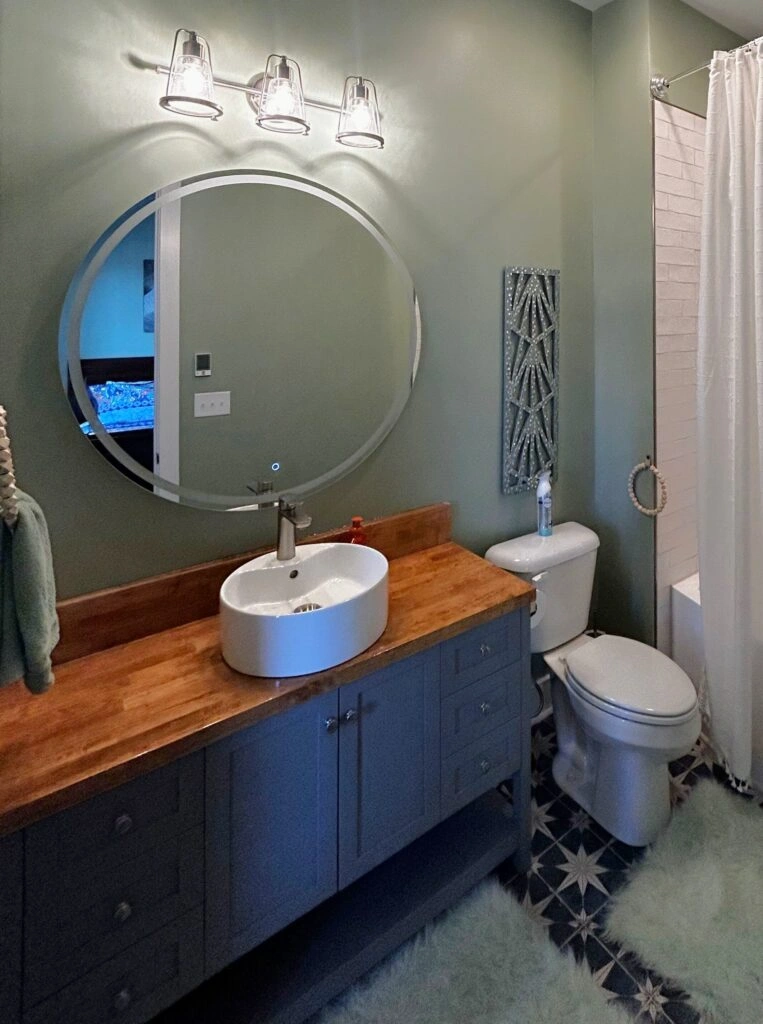 Bath & Bronze bathroom with blue vanity, wood countertop, round mirror, vessel sink, and vanity lighting.