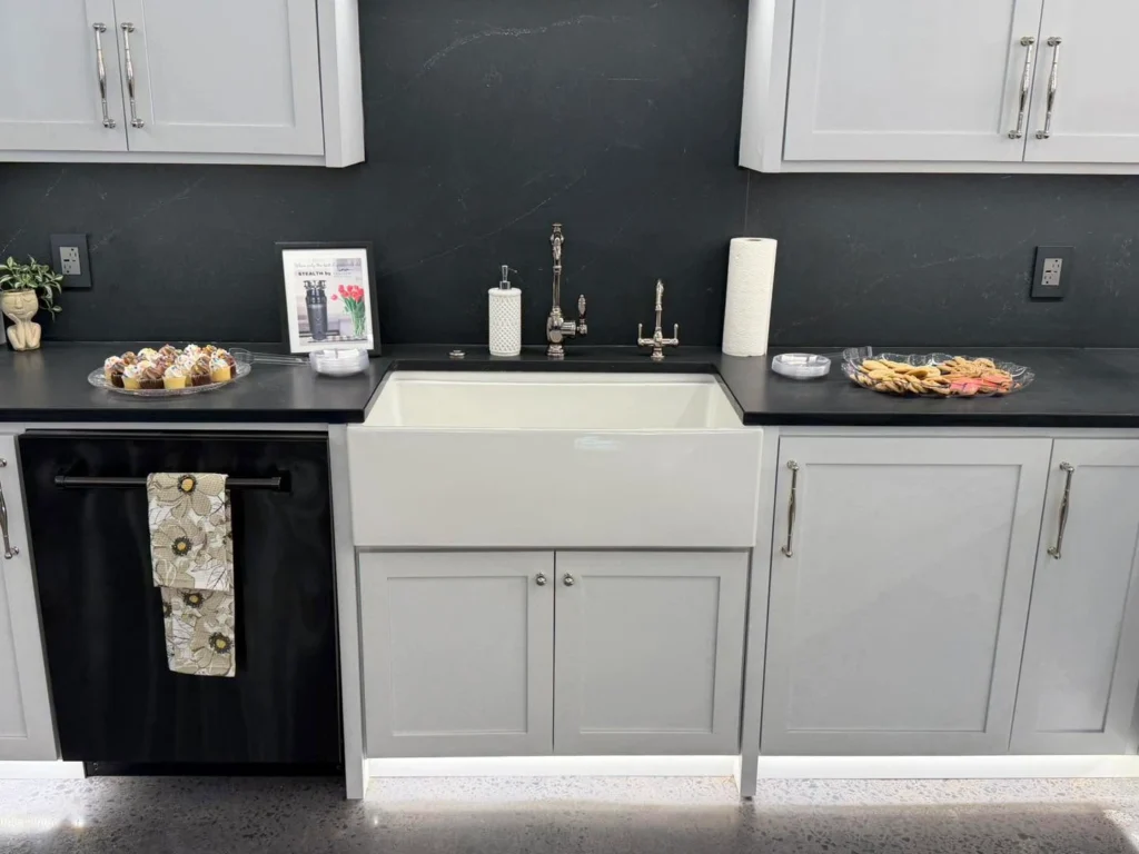 Kitchen showroom display with farmhouse sink, white cabinets, black countertop, and chrome fixtures.